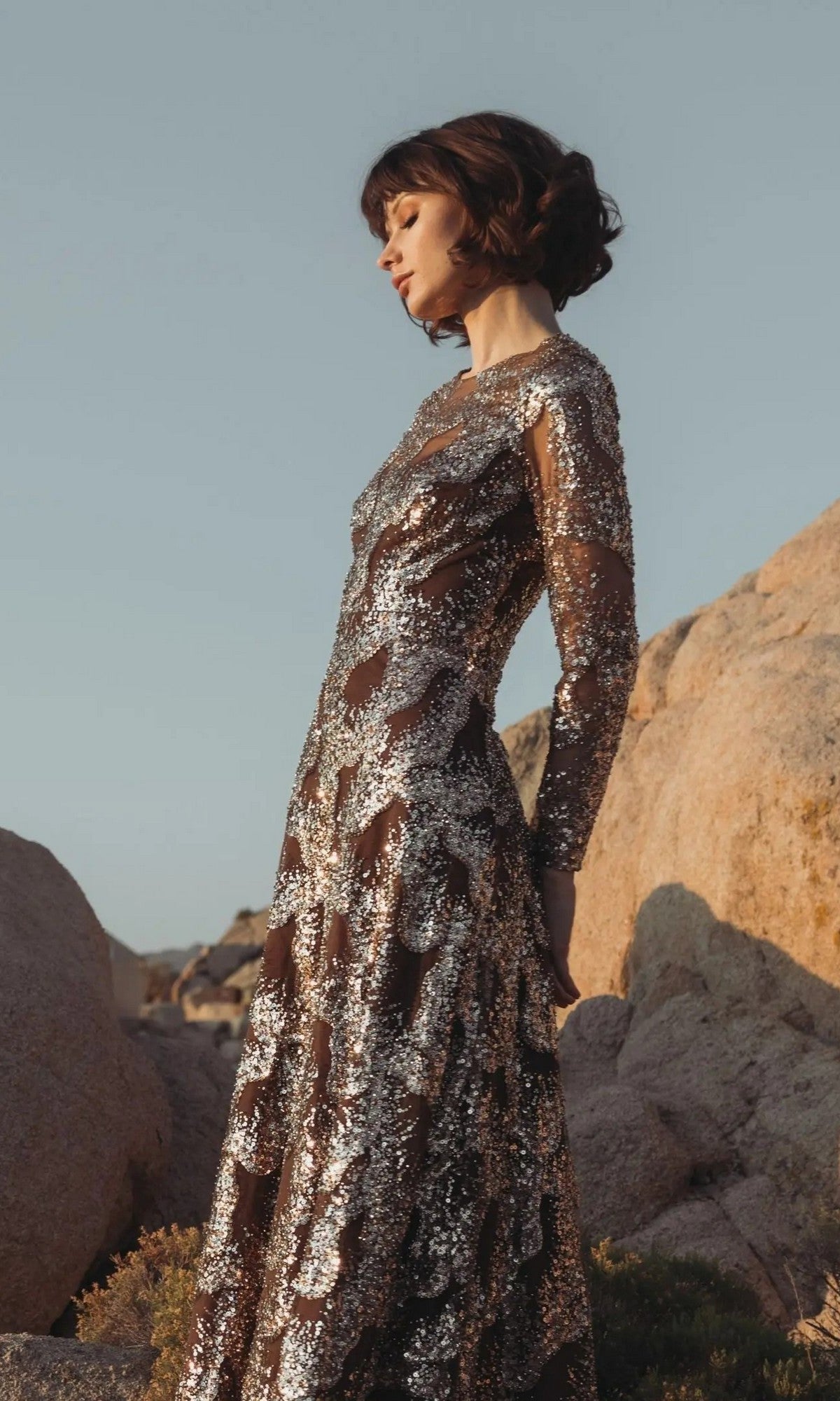 A woman wearing the Mac Duggal 69005 long formal dress, adorned with shimmering sequins and beads, stands among large rocks with her eyes closed, facing the sunlight.